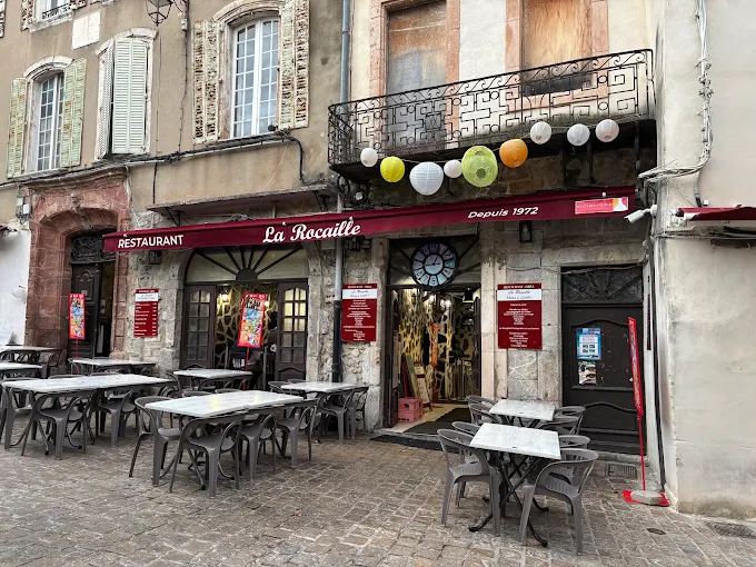 Les tables et chaises de La Rocaille dressées devant la façade rouge, sur la place pavée